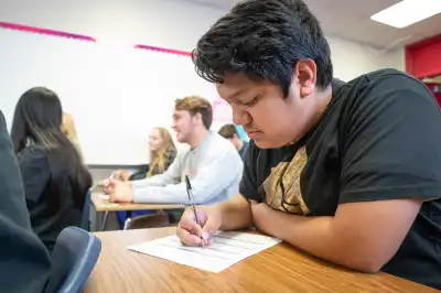 Junior Achievement high school student sitting at desk writing on paper in classroom with fellow students in background